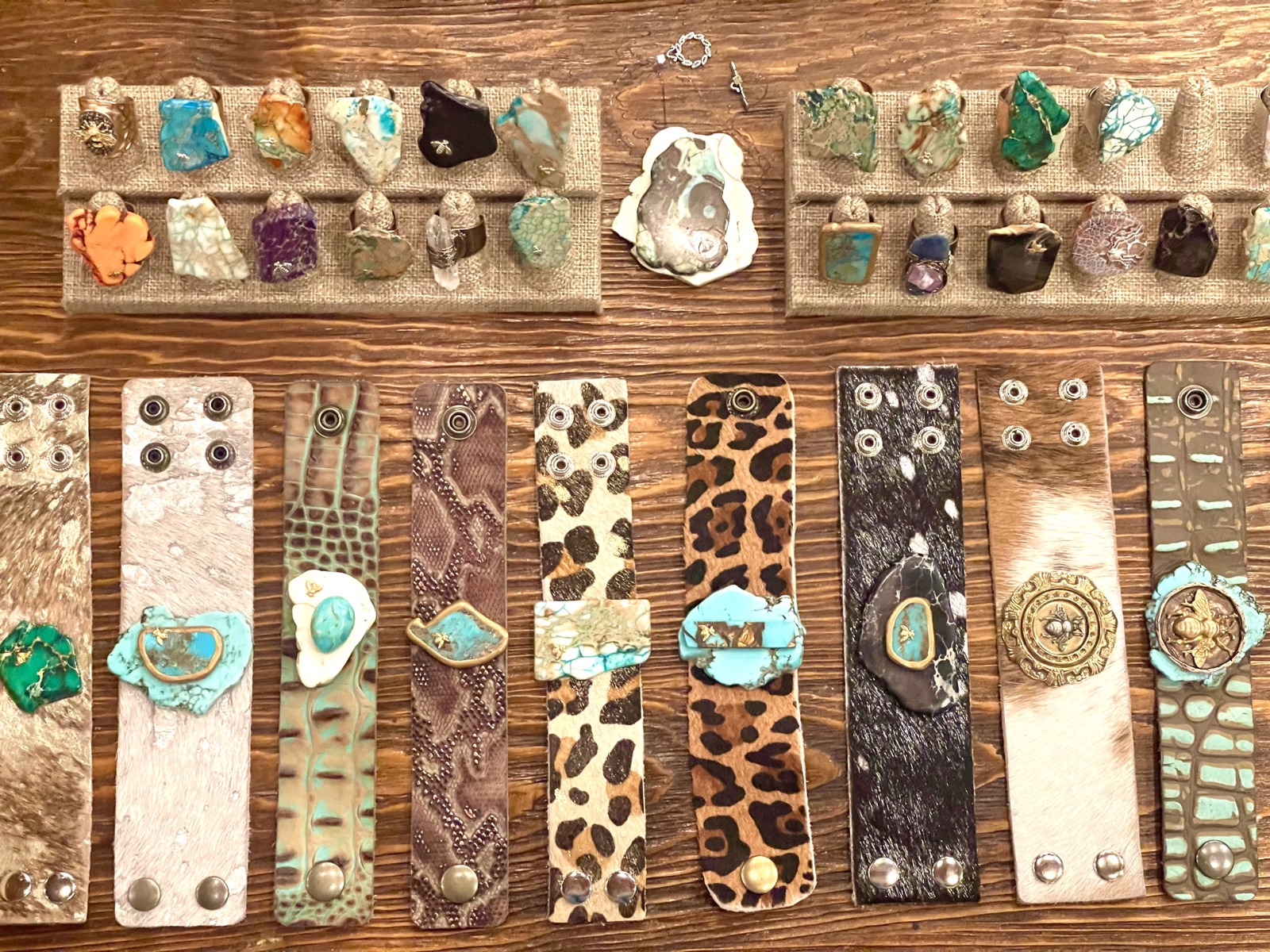 Two rows of stone rings on burlap above a line of leather and hide cuffs, each set with a stone or a medallion, on a wood table