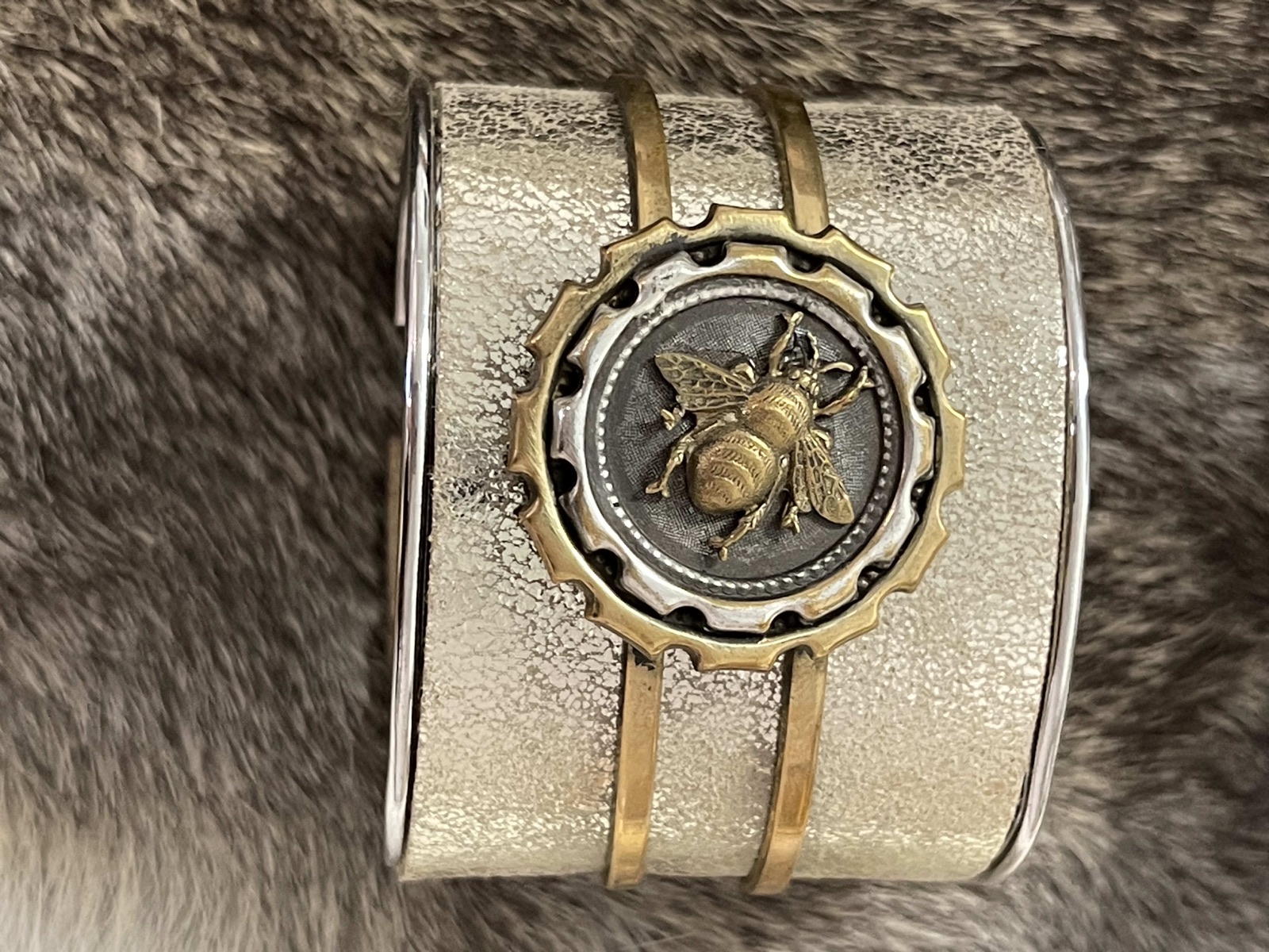 A wide silver-toned cuff with a brass bee medallion in a gear-shaped frame, on grey fur