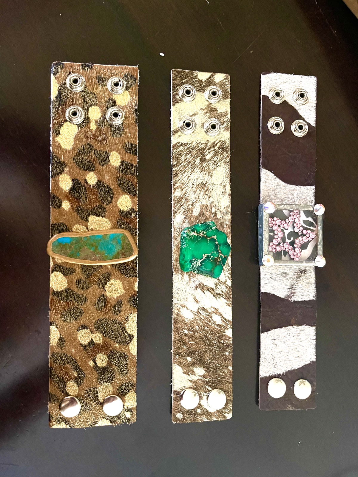 Three hide straps with snaps: gold-dotted leopard print with a long blue-green stone, speckled hide with a green stone, and zebra print with a pink-patterned square