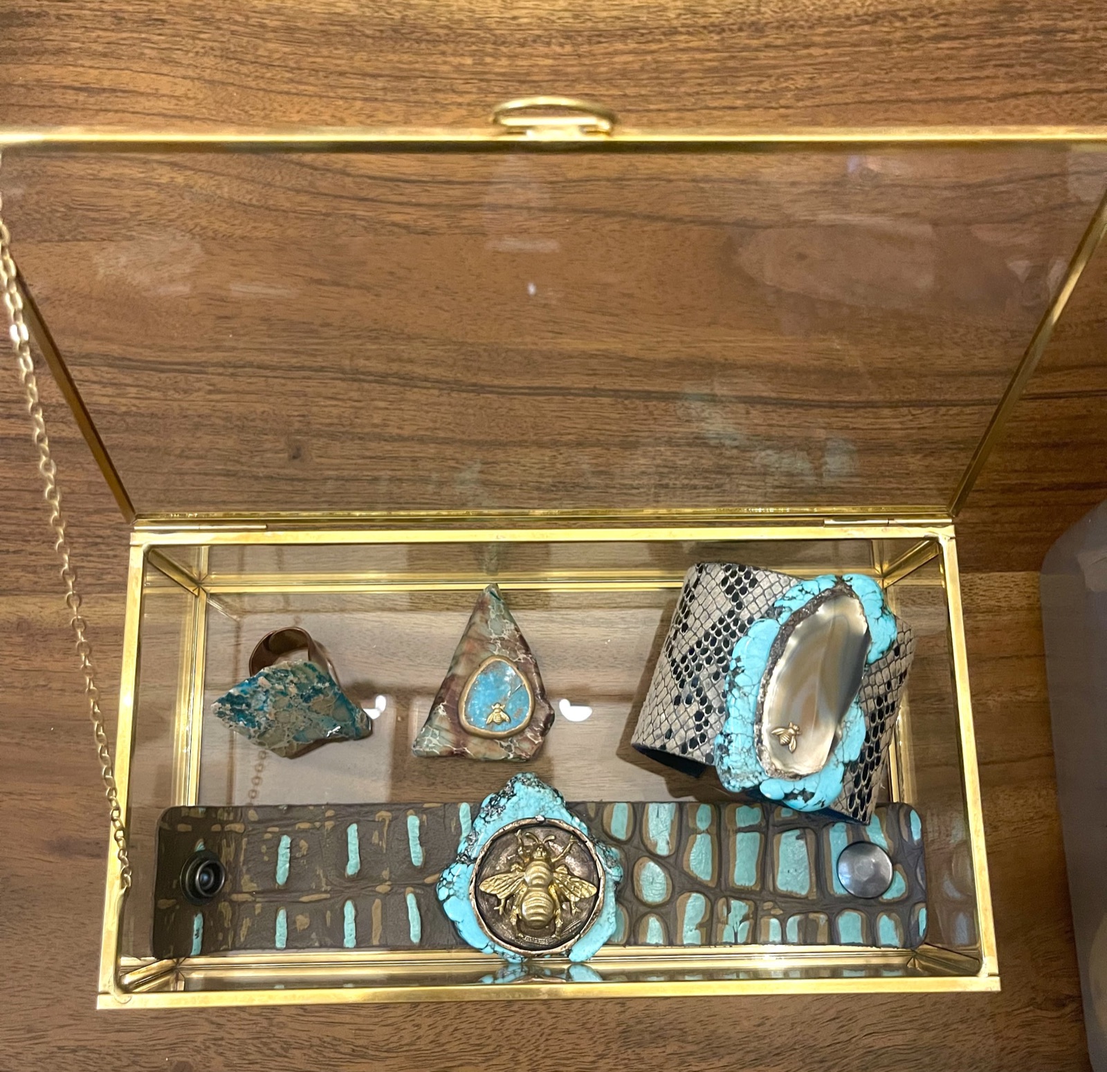 An open gold-framed glass box holding a crocodile-texture cuff with a bee medallion ringed in blue-green, a snakeskin-texture cuff and two stone pendants