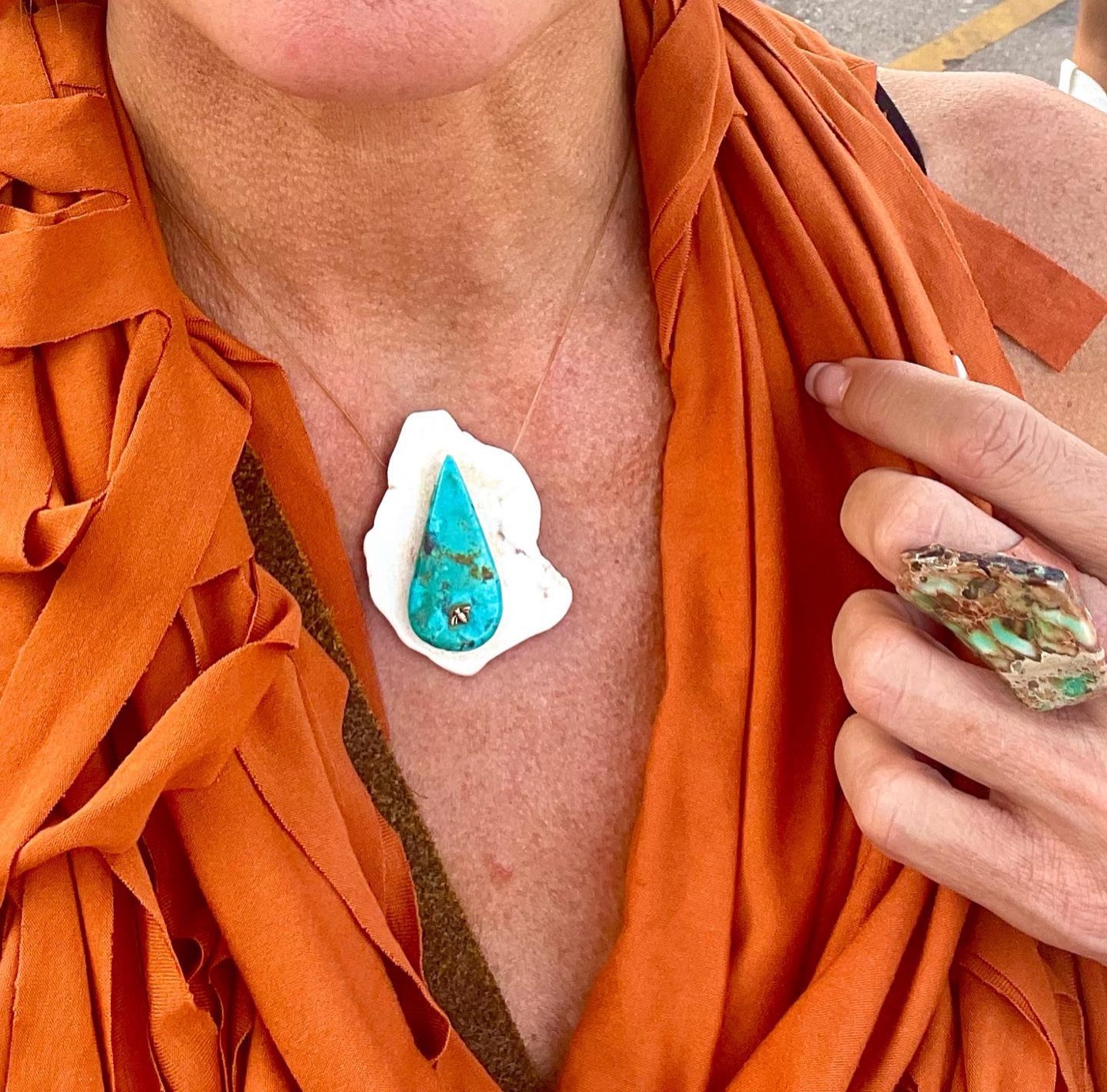 A turquoise teardrop pendant with a white stone rim on an orange draped top, a ring on the hand