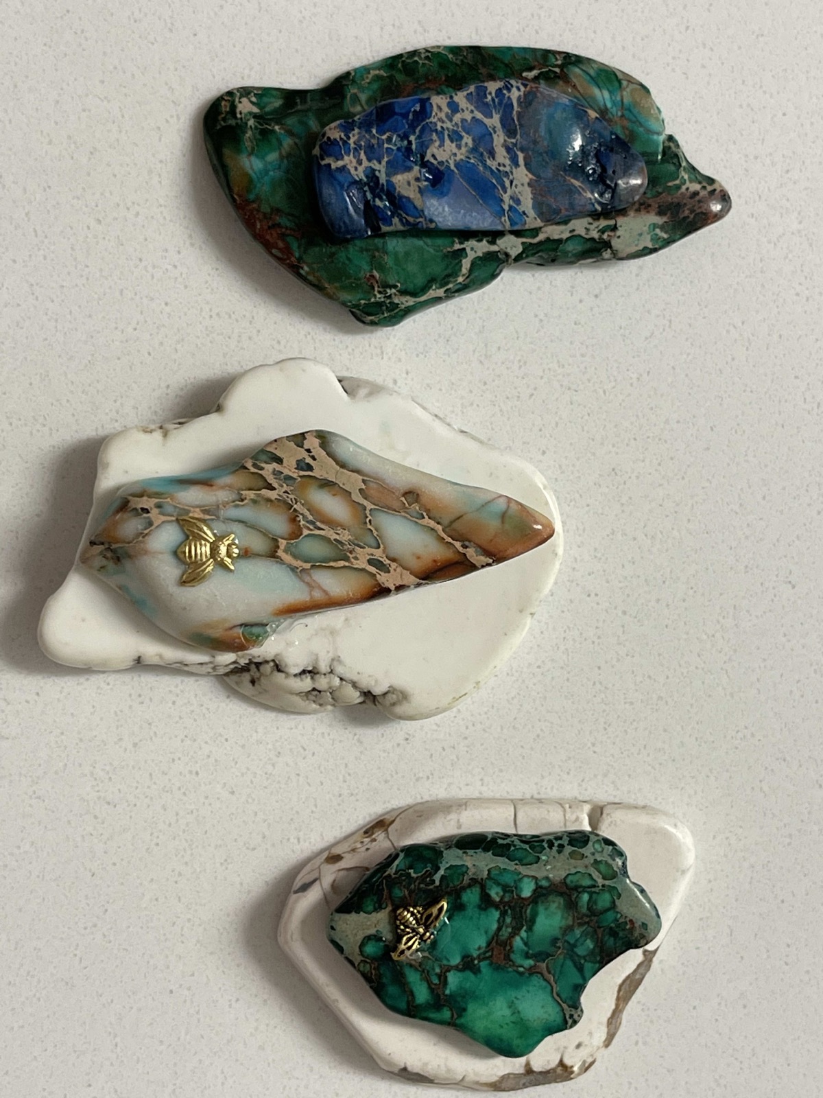 Three stone pendants on a white backdrop: a blue and green stone, a pale stone in a white rim with a small gold bee, and a green stone in a white rim with a small gold bee