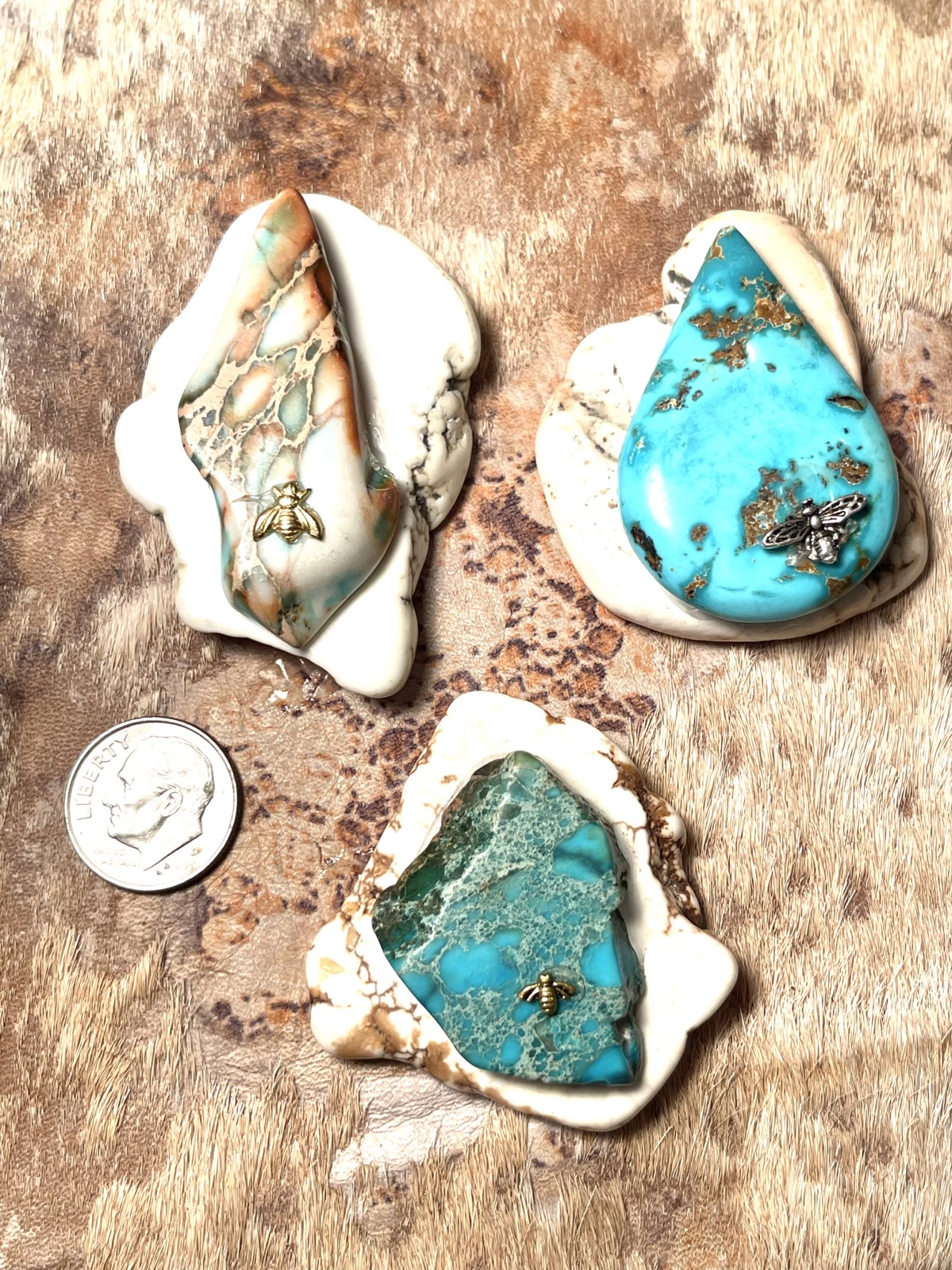 Three stone pendants with white rims and small bees, beside a dime for scale, on a speckled hide