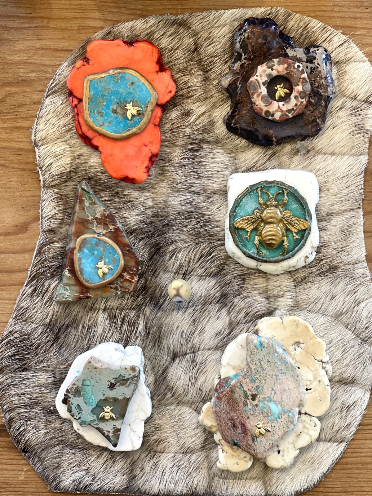 Six stone pendants with small gold bee castings laid out on grey fur, in orange, brown, blue-green and cream